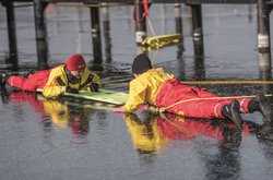 Ein liegender Rettungsschwimmer zieht ein anderes DLRG-Mitglied bei einer Übung mit einem langen Holzbrett aus einem Loch im Eis.