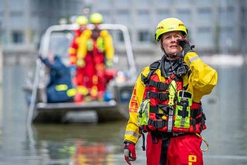 Person in rot-gelber Einsatzkleidung rechts im Vordergrund, im Hintergrund weitere Personen in Einsatzkleidung auf Boot im Wasser